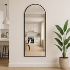 Full-length mirror with black frame leaning against a wall in a room with a plant and kitchen reflection.