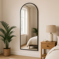 Full-length mirror leaning against a wall in a bedroom with a plant and nightstand.