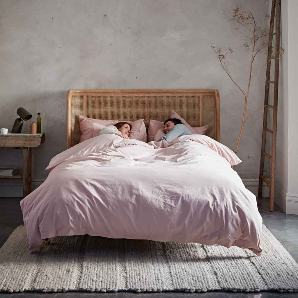 Two people lying in bed under a pink duvet in a bedroom setting.