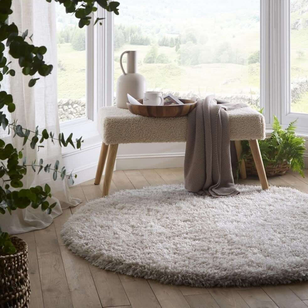 Round fluffy rug in a room with a stool, plants, and a large window.