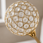 A close-up of a decorative crystal globe lampshade with a gold metal frame and a gold finial stem, set against a softly blurred neutral background.