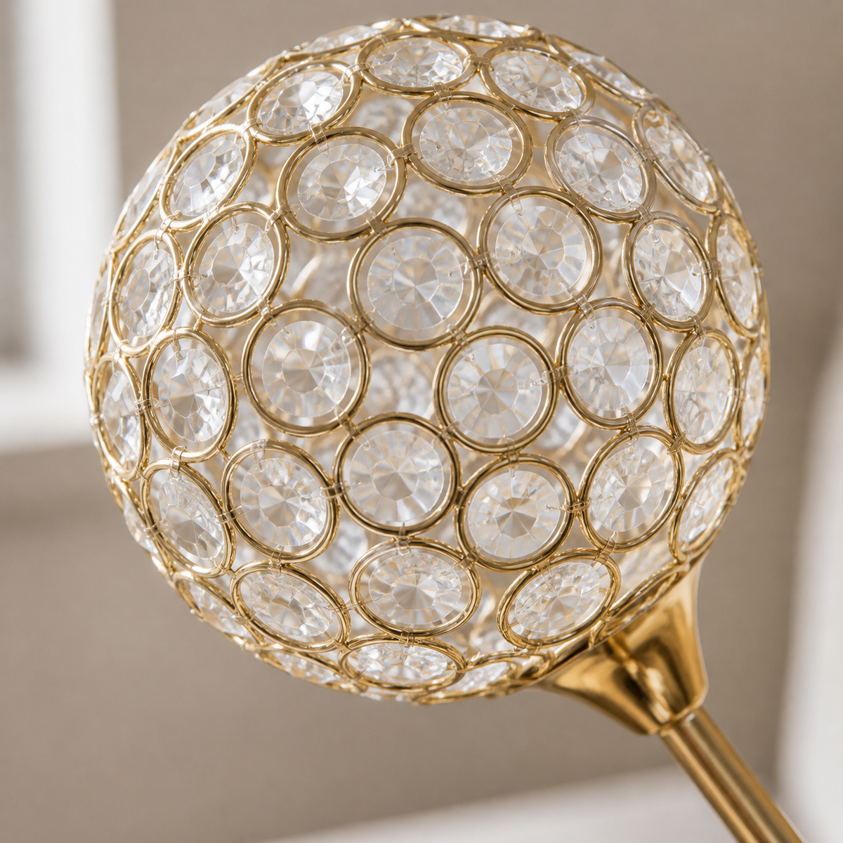 A close-up of a decorative crystal globe lampshade with a gold metal frame and a gold finial stem, set against a softly blurred neutral background.