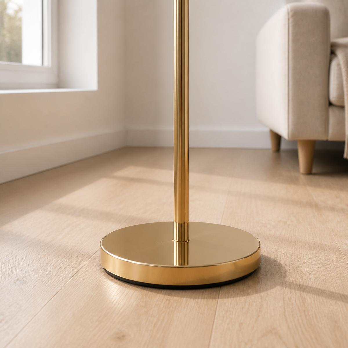A close-up of a polished gold floor lamp base and pole standing on a light wood floor in a bright living room with a beige armchair and window in the background.