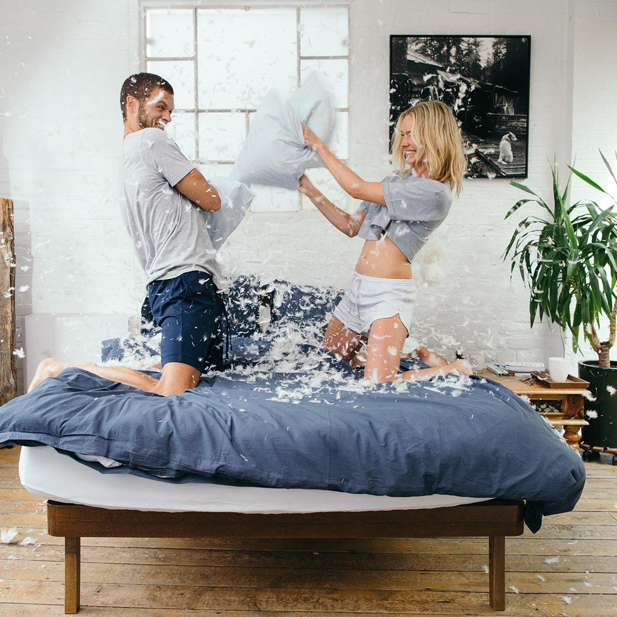 Two people playing with pillows on a bed in a bright room.