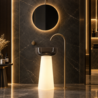A modern bathroom scene featuring a freestanding pedestal sink with a glowing illuminated base, a curved matte brass faucet, a round backlit wall mirror, and dark marble walls and floor.
