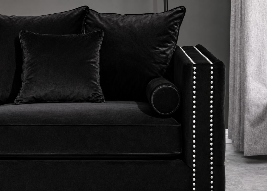 A close-up of a black velvet sofa with matching black throw pillows, featuring a cylindrical bolster pillow and a black armrest accented with white nailhead trim, set against a dark gray wall and light gray curtain.