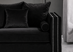 A close-up of a black velvet sofa with matching black throw pillows, featuring a cylindrical bolster pillow and a black armrest accented with white nailhead trim, set against a dark gray wall and light gray curtain.