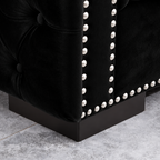 Close-up of a black tufted velvet upholstered furniture corner with silver nailhead trim and a dark wood base on a gray tiled floor.