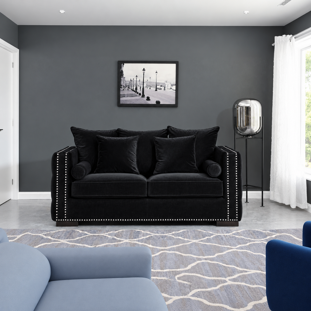 A black velvet loveseat sofa with silver nailhead trim, styled in a modern living room with a gray wall, framed black-and-white artwork, a floor lamp, and a blue patterned rug.