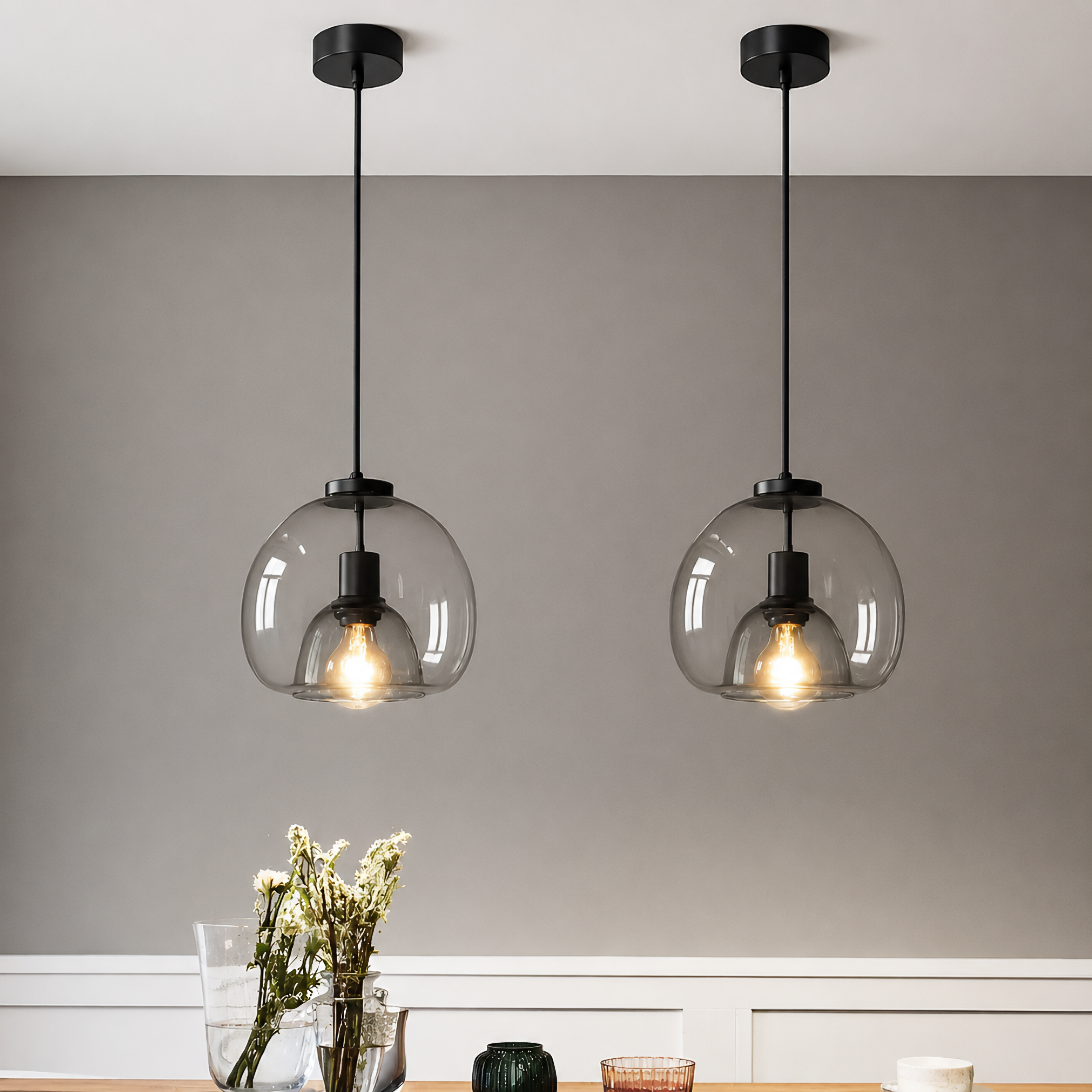Two matching black pendant lights with large round clear glass shades hang symmetrically against a gray wall above a white wainscoted table. Warm exposed light bulbs are visible inside the glass globes, and part of a vase with dried stems and table decor appear at the bottom.