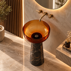 An amber glass bowl sink with a dark marble pedestal base, set in a modern bathroom with warm lighting and marble tiles.