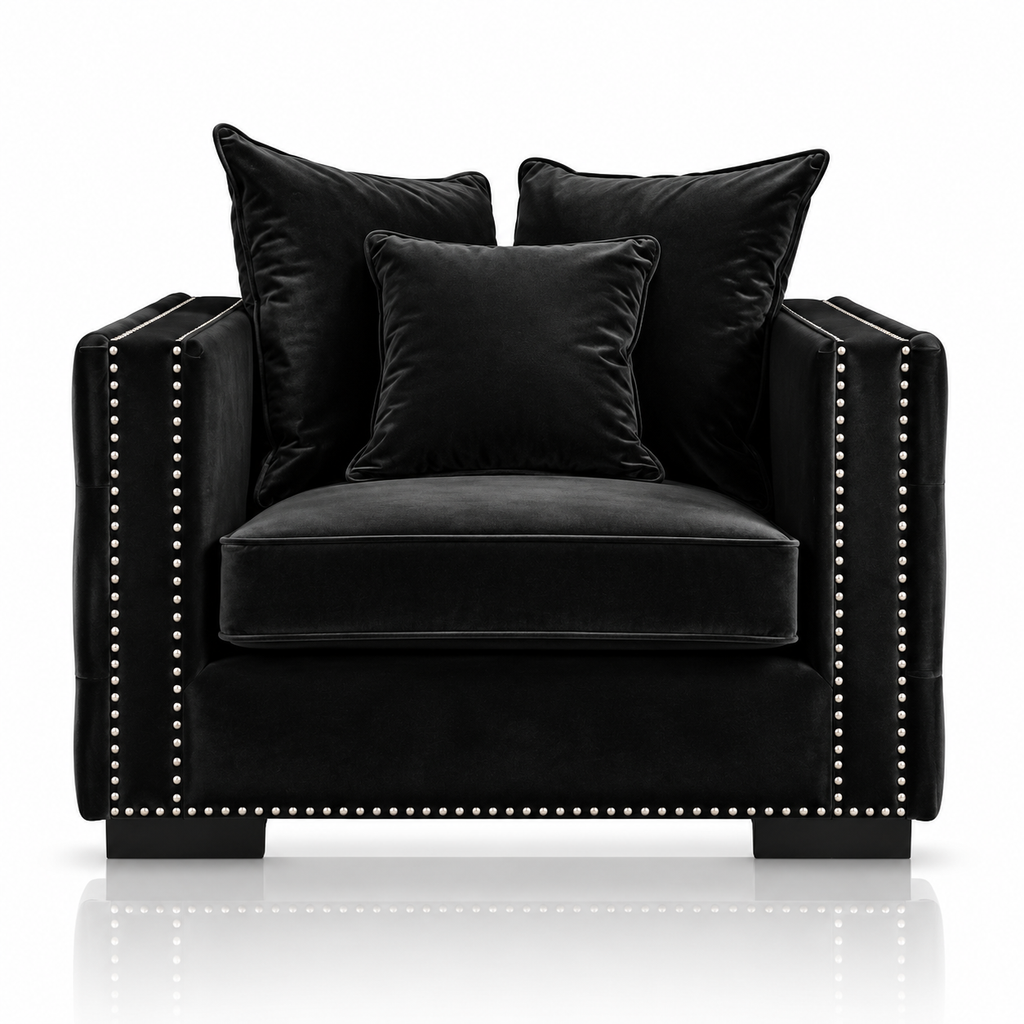 A black velvet armchair with matching throw pillows and nailhead trim, photographed on a clean white background with a subtle reflection below.
