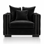 A black velvet armchair with matching throw pillows and nailhead trim, photographed on a clean white background with a subtle reflection below.