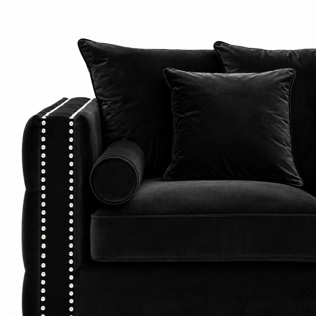 Black velvet sofa with matching throw pillows and a cylindrical bolster pillow, featuring silver nailhead trim on the armrest, shown against a white background.