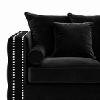 Black velvet sofa with matching throw pillows and a cylindrical bolster pillow, featuring silver nailhead trim on the armrest, shown against a white background.