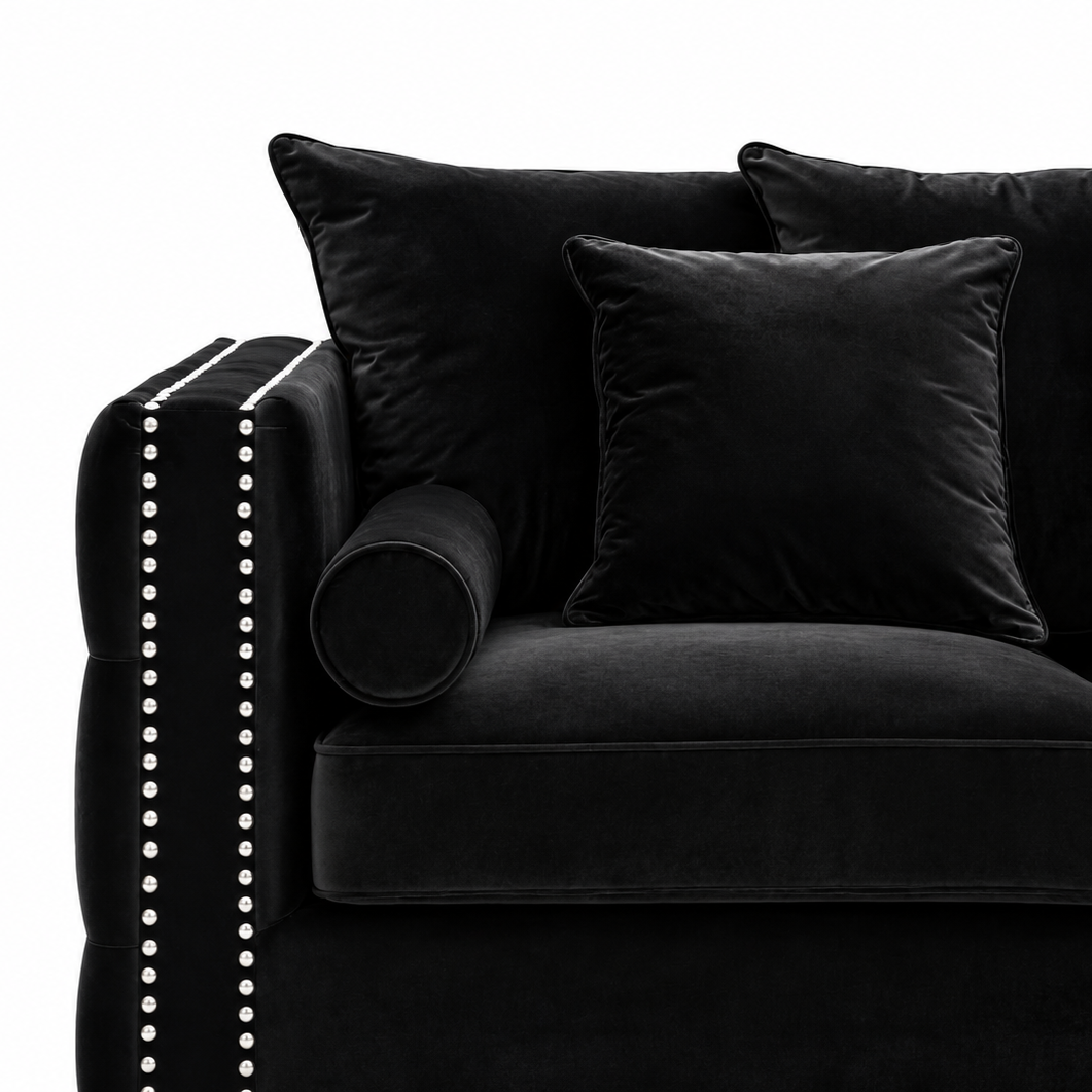 Black velvet sofa with matching throw pillows and a cylindrical bolster pillow, featuring silver nailhead trim on the armrest, shown against a white background.
