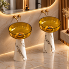 Two modern pedestal sinks with translucent amber glass bowls and white marble bases with black veining, set in a luxurious bathroom with a large illuminated mirror and gold fixtures.