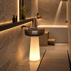 A modern bathroom scene featuring a glowing illuminated pedestal sink with a dark glass bowl, set against warm stone tile walls and floor. A bathtub with a shower head is visible in the background, along with a dark shelf, bottles, and a bath mat.