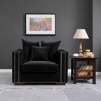 A black velvet armchair with silver nailhead trim is styled in a modern living room setting against a dark gray wall, featuring a framed landscape print, a wooden side table with a lamp, and a gray rug.