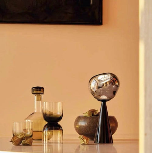 Decorative items including a bottle, glasses, and a metallic bowl on a reflective surface with a neutral background.