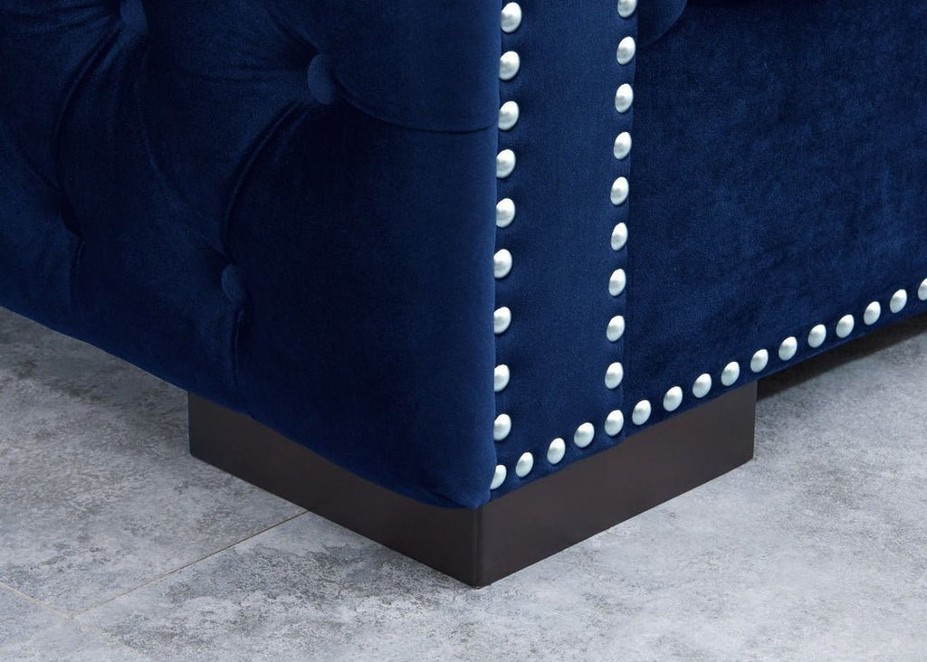 Close-up of a blue velvet sofa with nailhead trim on a gray floor.