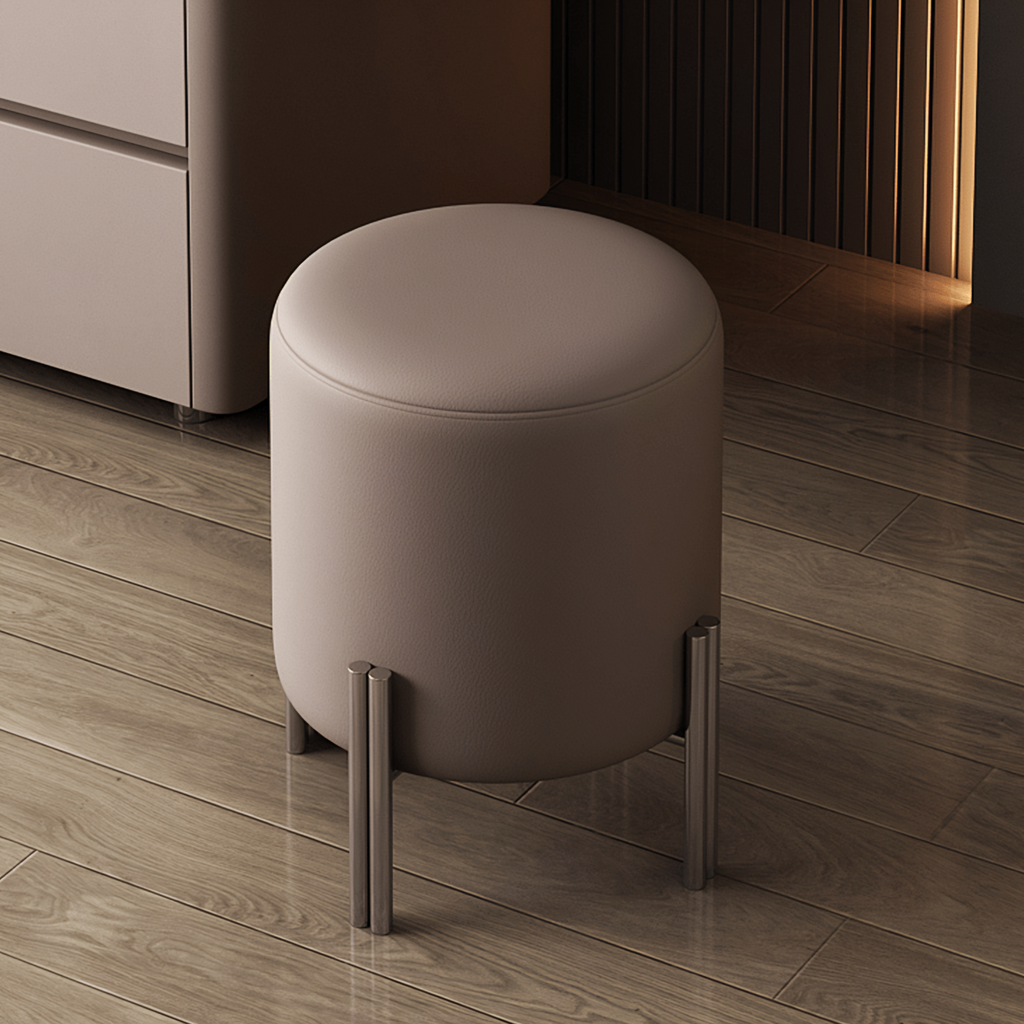 A cylindrical upholstered stool with a light taupe cushion and slim metallic legs, shown in a modern interior with wood flooring and furniture in the background.