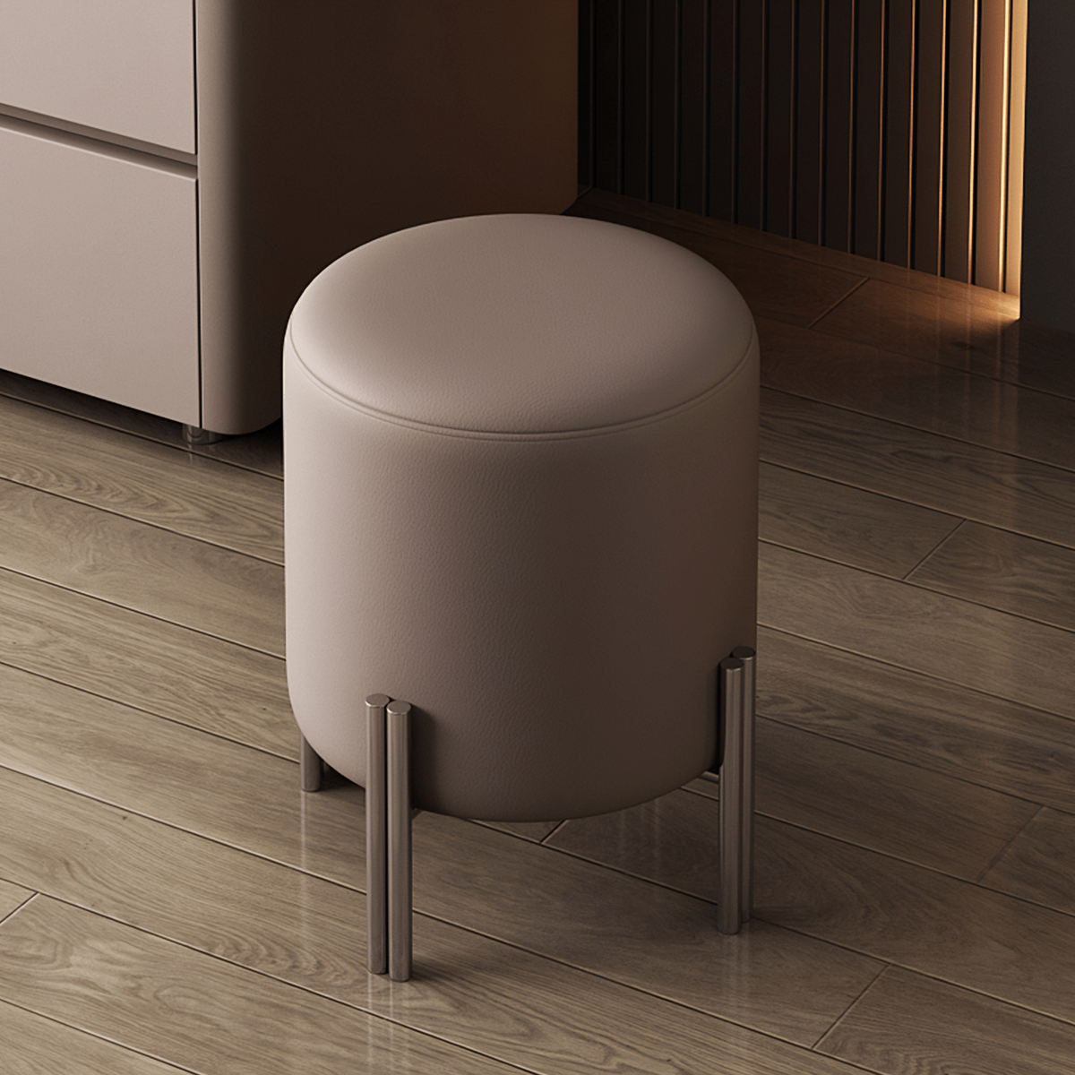 A cylindrical upholstered stool with a light taupe cushion and slim metallic legs, shown in a modern interior with wood flooring and furniture in the background.