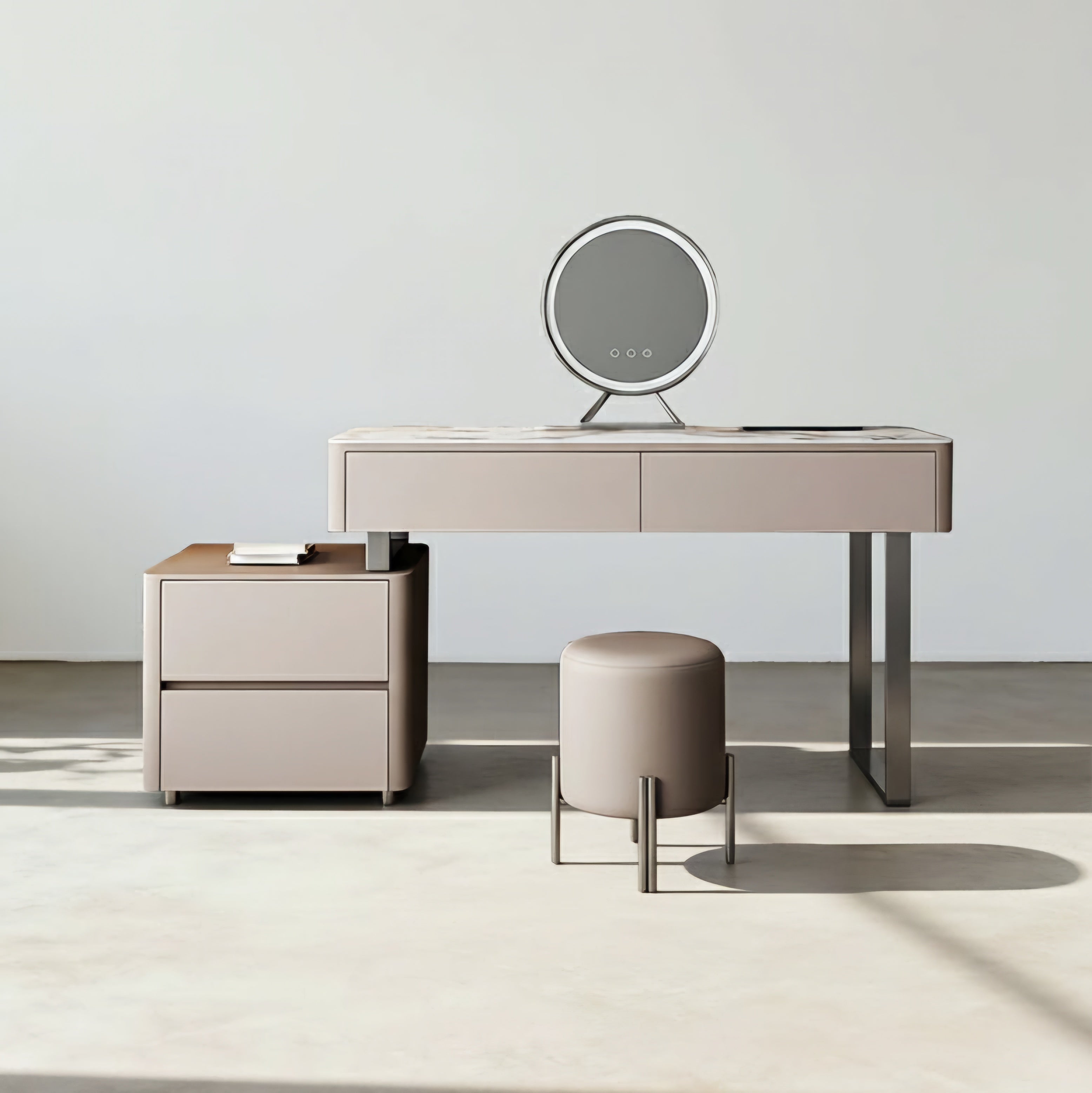 A modern vanity desk set in a minimalist studio, featuring a light beige desk with a marble-look top, a matching two-drawer nightstand, a round upholstered stool on metal legs, and an oval LED mirror.