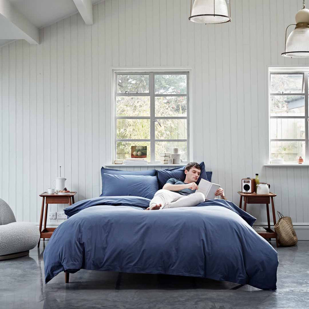 Person lying on a bed with blue bedding in a bright bedroom.