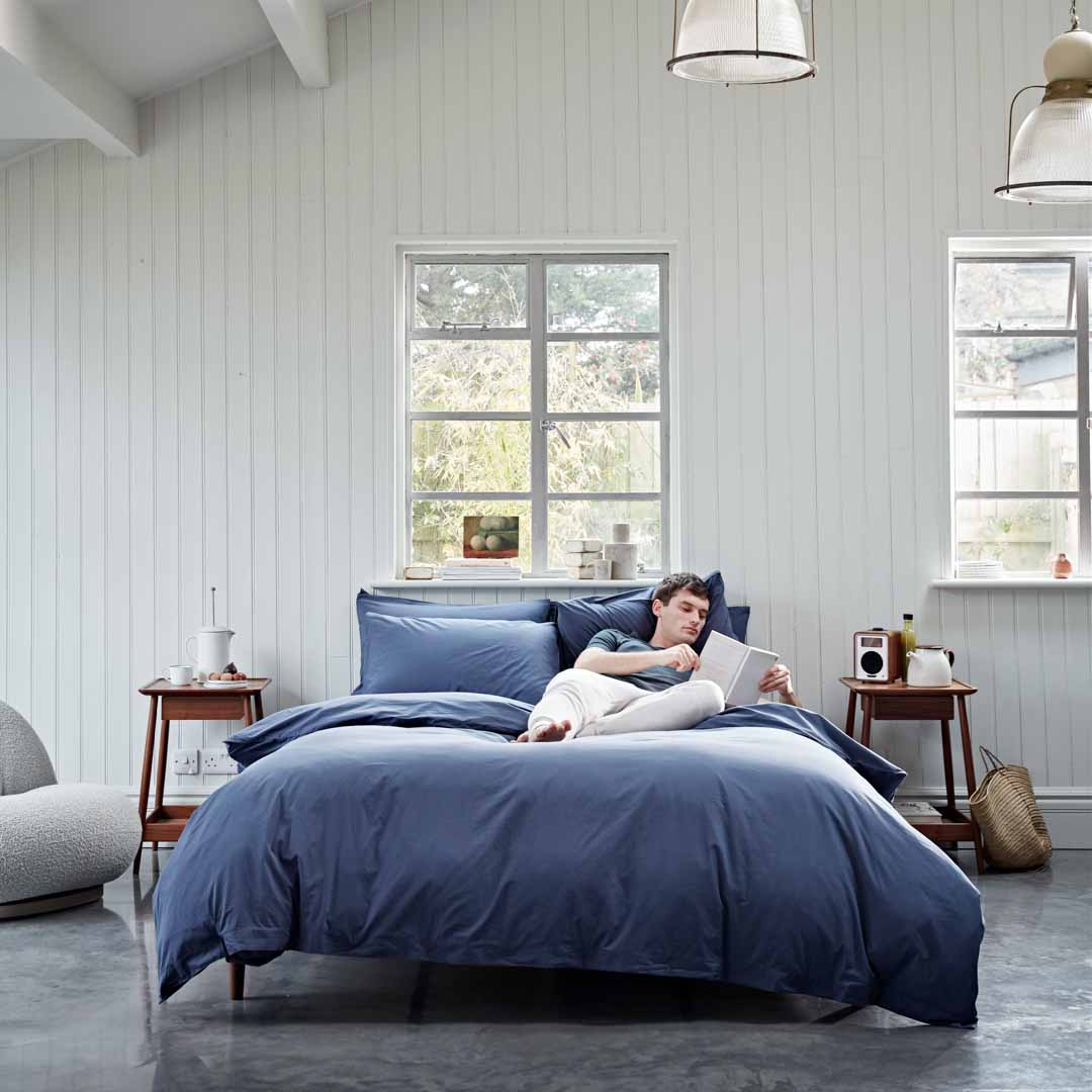 Person lying on a bed with blue bedding in a bright bedroom.