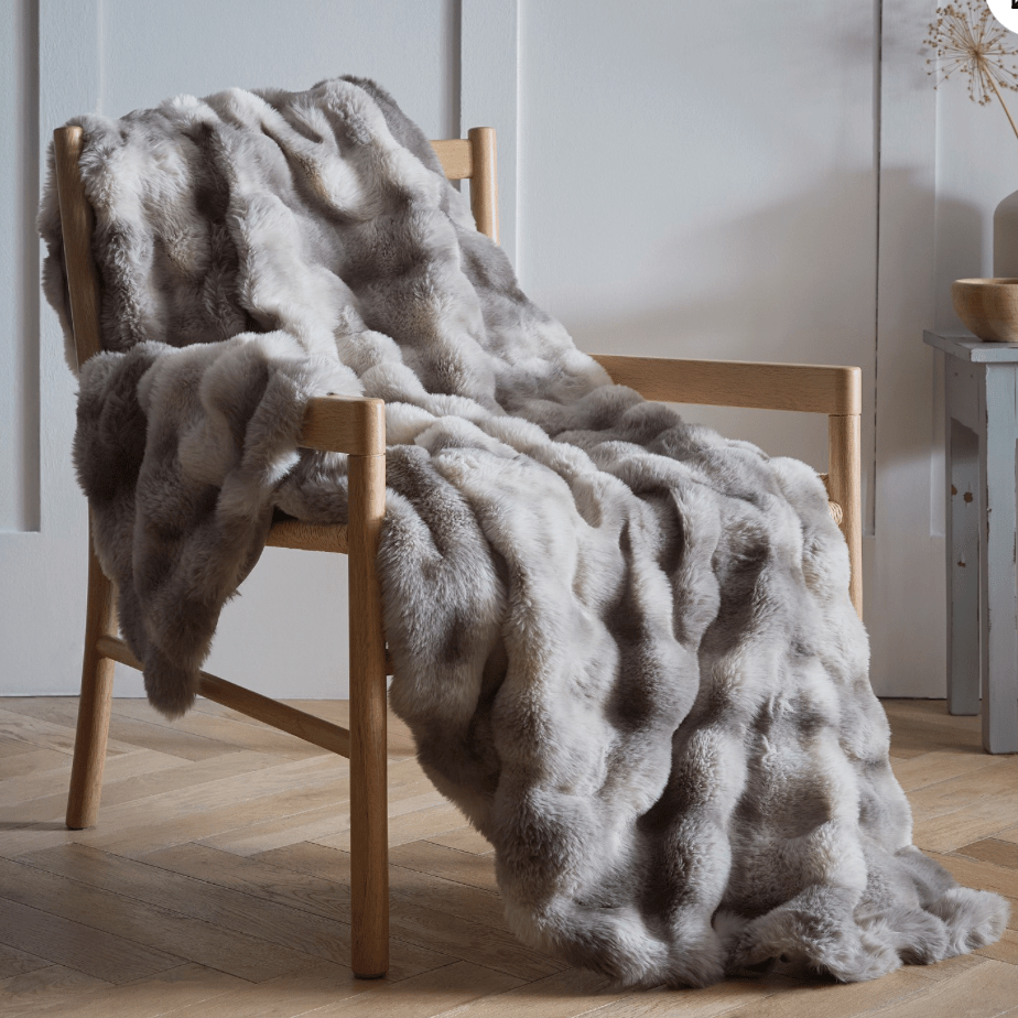 Gray fur blanket draped over a wooden chair in a room with a light-colored wall and wooden floor.