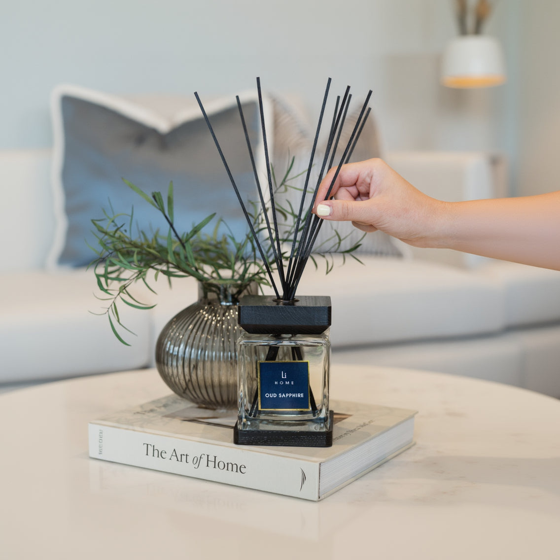 Hand placing diffuser sticks into a diffuser bottle labeled 'U home' on a book with a plant in the background.