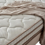 A close-up of a quilted mattress with a textured cream-colored cover, styled with a beige and white patterned throw blanket draped across the top.