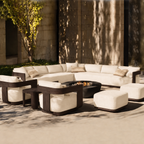 Outdoor patio furniture set with a curved sectional sofa, two armchairs, two ottomans, and a round black fire pit table, arranged on a stone patio in front of a modern building wall with tree shadows.