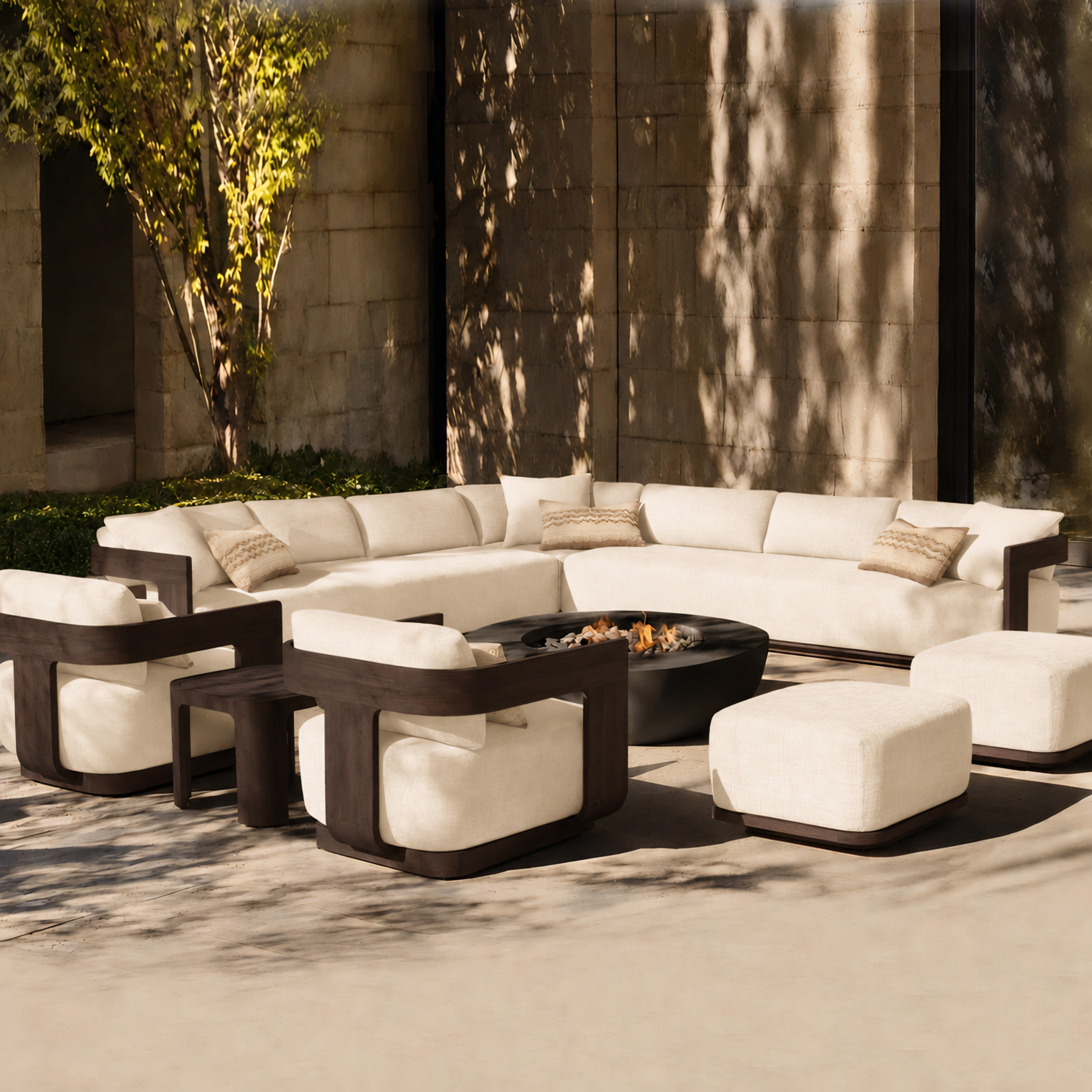 Outdoor patio furniture set with a curved sectional sofa, two armchairs, two ottomans, and a round black fire pit table, arranged on a stone patio in front of a modern building wall with tree shadows.