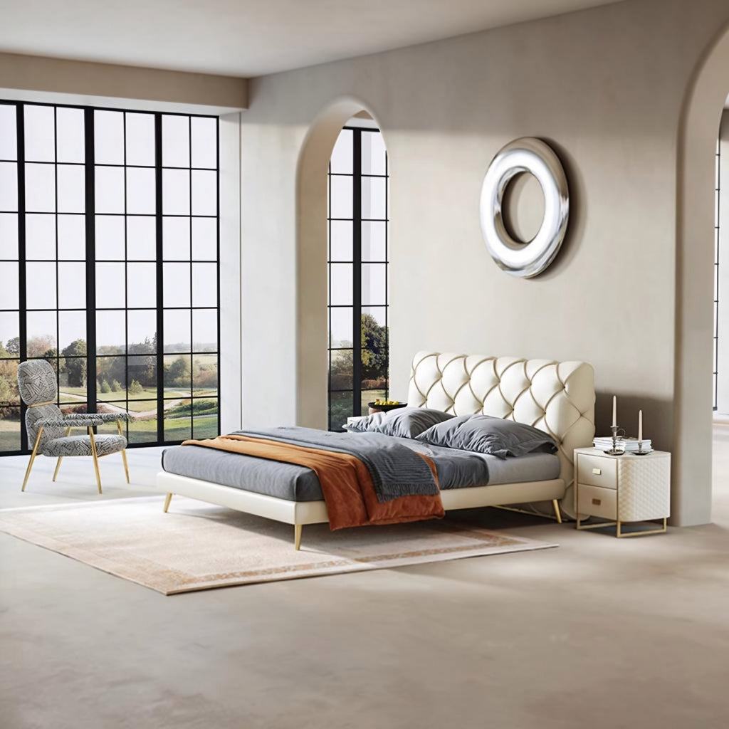 A modern cream tufted platform bed with gold legs, styled with gray bedding and an orange throw blanket, set in a minimalist beige bedroom with a matching nightstand and large window views.