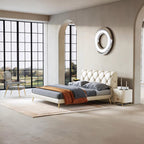 A modern cream tufted platform bed with gold legs, styled with gray bedding and an orange throw blanket, set in a minimalist beige bedroom with a matching nightstand and large window views.