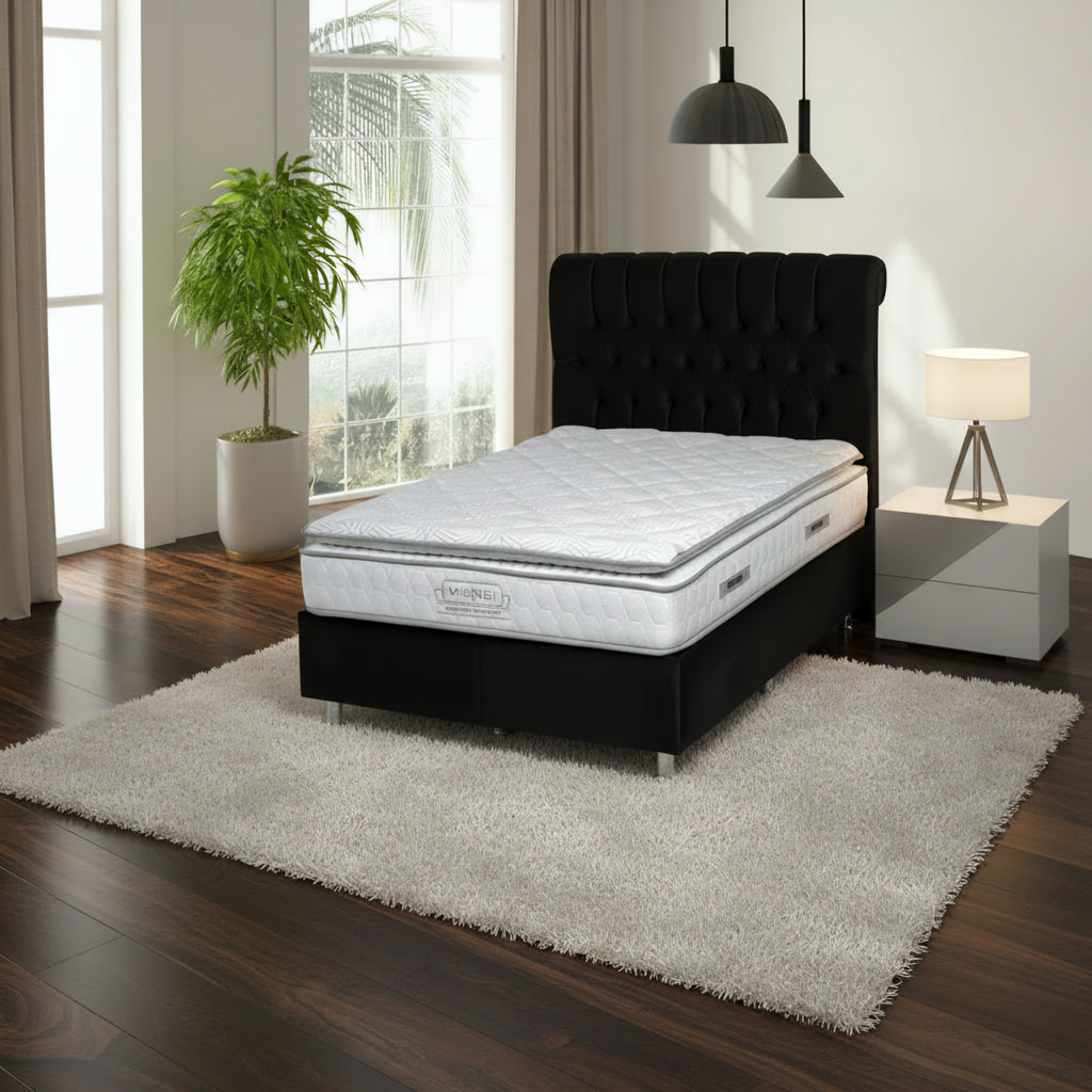 A dark gray tufted bed frame with a white quilted mattress, shown in a bright bedroom setting with a window, potted plant, and pendant lights.