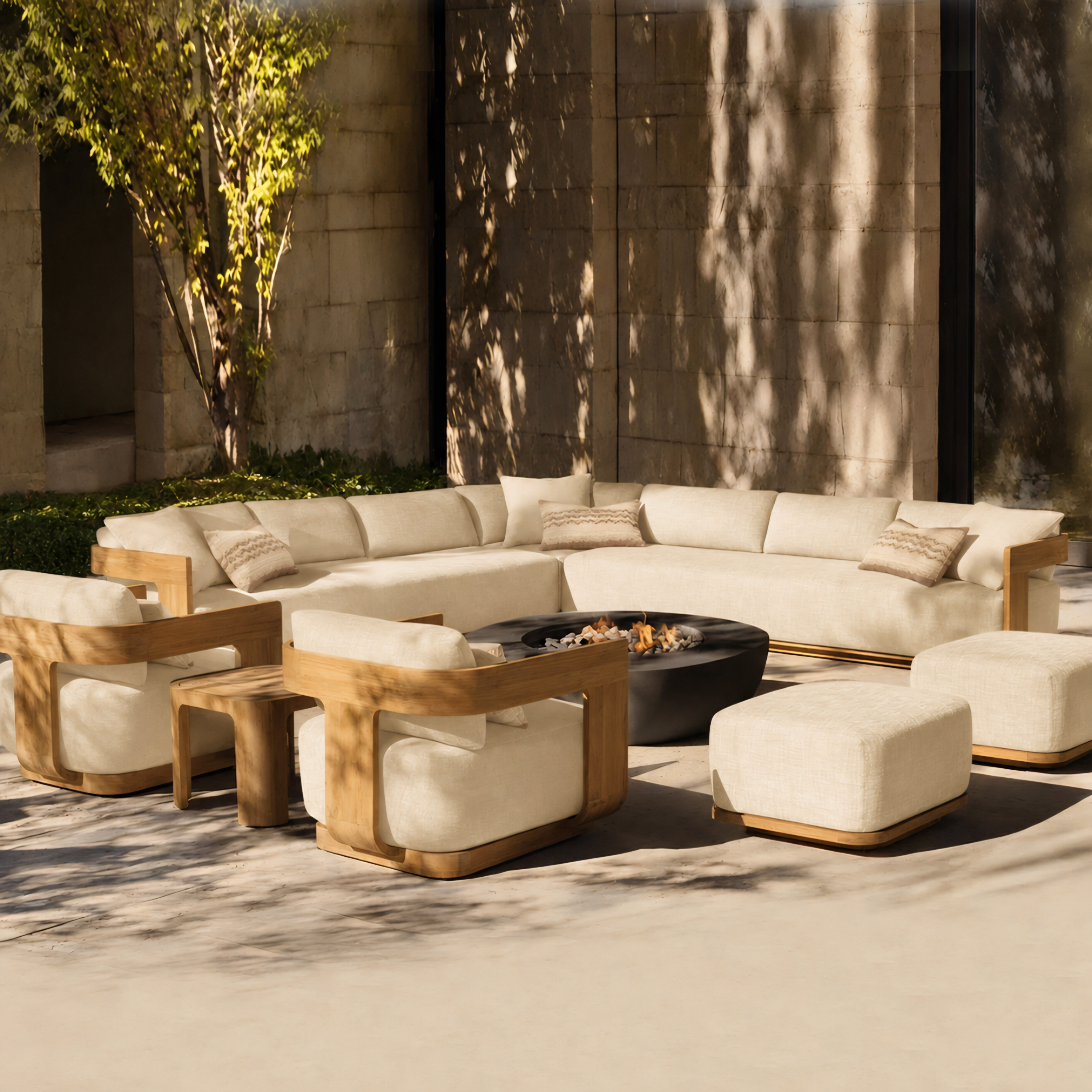 Outdoor patio furniture set with cream upholstered sectional chairs, matching ottomans, two wooden armchairs, and a round black fire pit table, arranged in front of a stone wall with dappled sunlight.