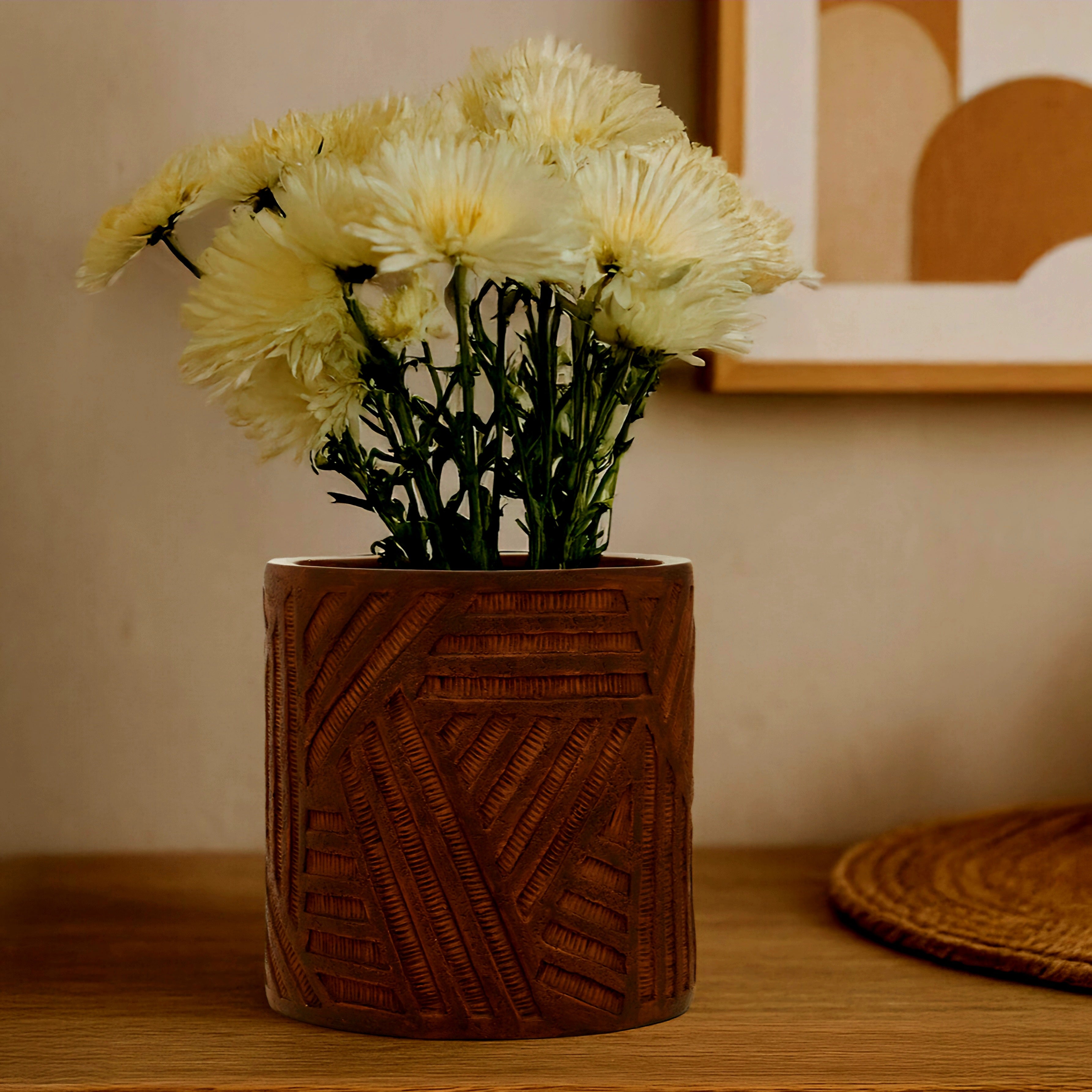 A brown textured ceramic pot with carved geometric patterns, filled with pale yellow chrysanthemums, sitting on a wooden table in a warm, neutral-toned room.