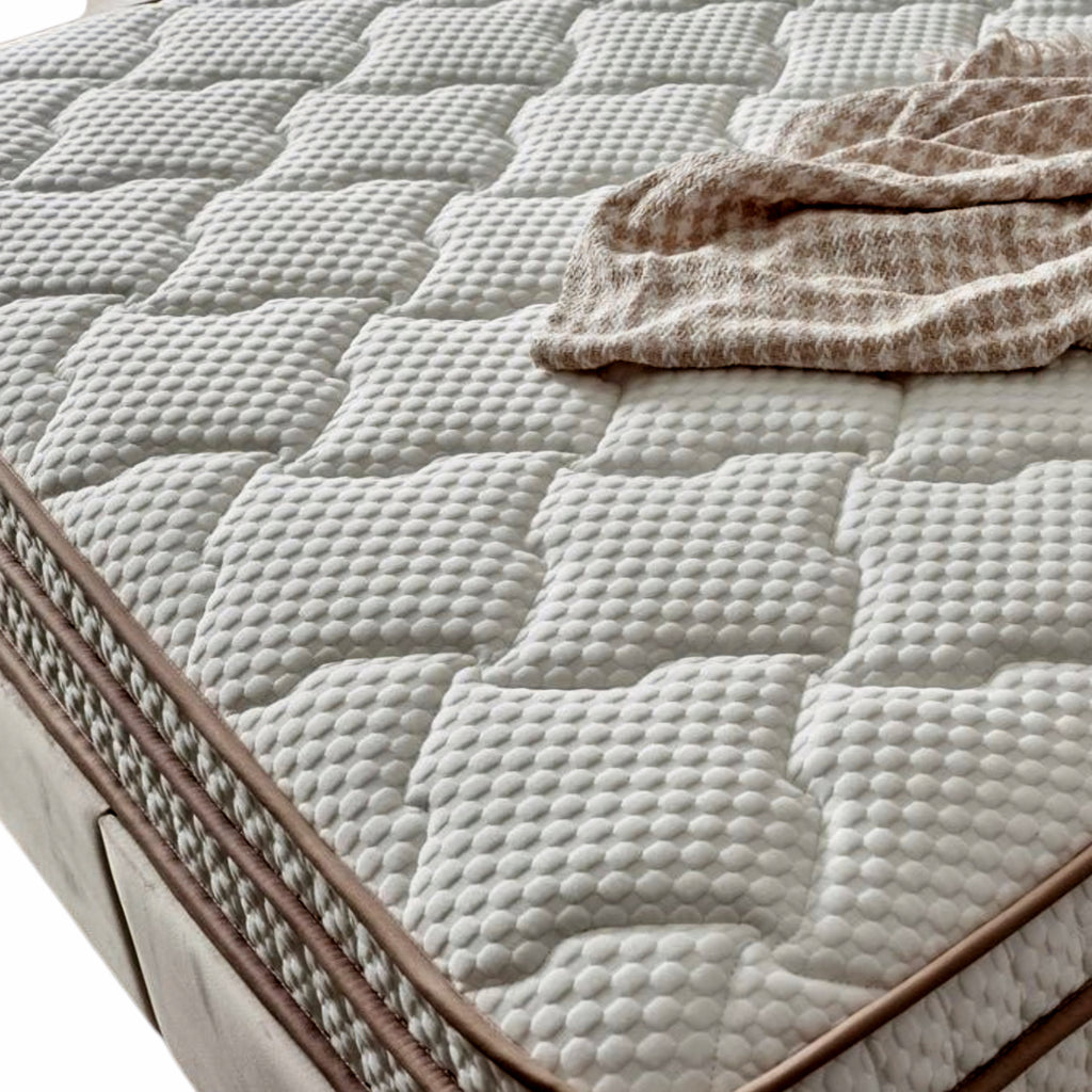 A close-up of a quilted mattress topper or mattress surface with a textured diamond pattern, brown trim, and a beige patterned throw blanket draped on top.
