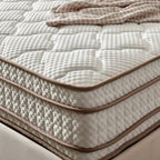 A close-up of a quilted mattress with a textured white top surface and side panels, finished with tan piping, with a beige patterned throw blanket draped across the upper corner.