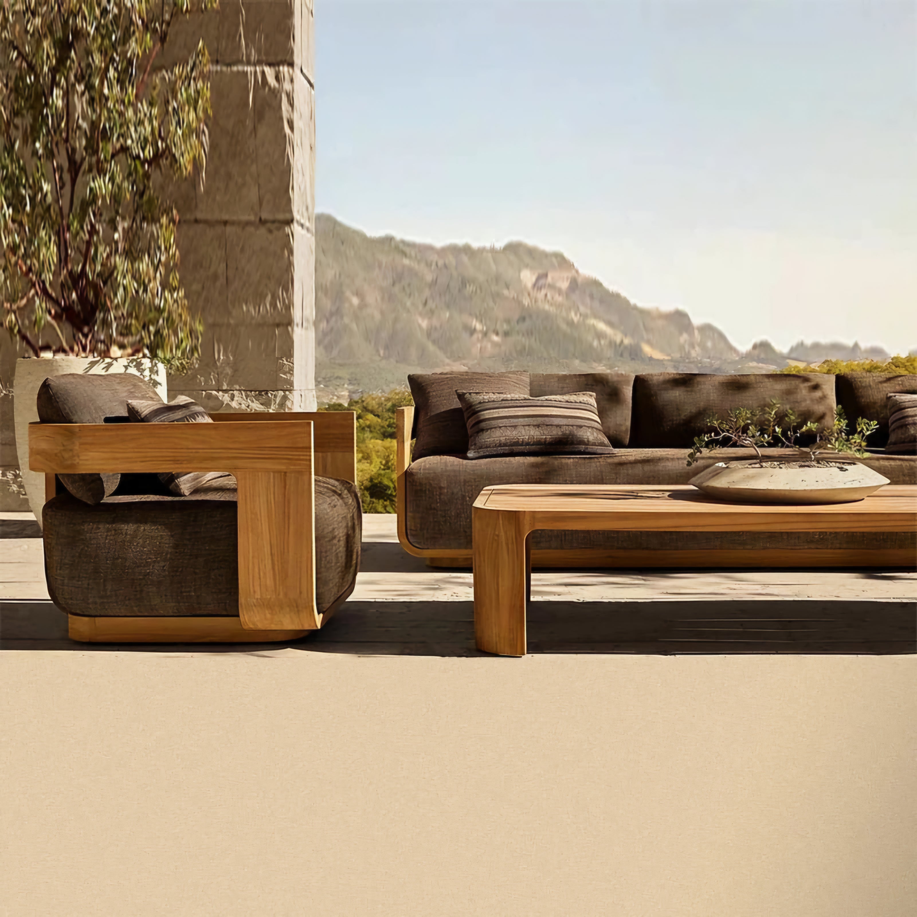 Modern outdoor patio furniture set with a wooden armchair, matching coffee table, and a dark brown sectional sofa, styled on a concrete patio with a stone column and mountain backdrop.