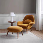 Mustard yellow armchair and ottoman with a white lamp on a wooden side table in a room with gray walls and white baseboards.