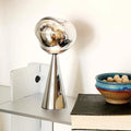 Modern silver lamp on a white surface with a book and decorative bowl in the background.