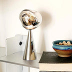 Modern silver lamp on a white surface with a book and decorative bowl in the background.