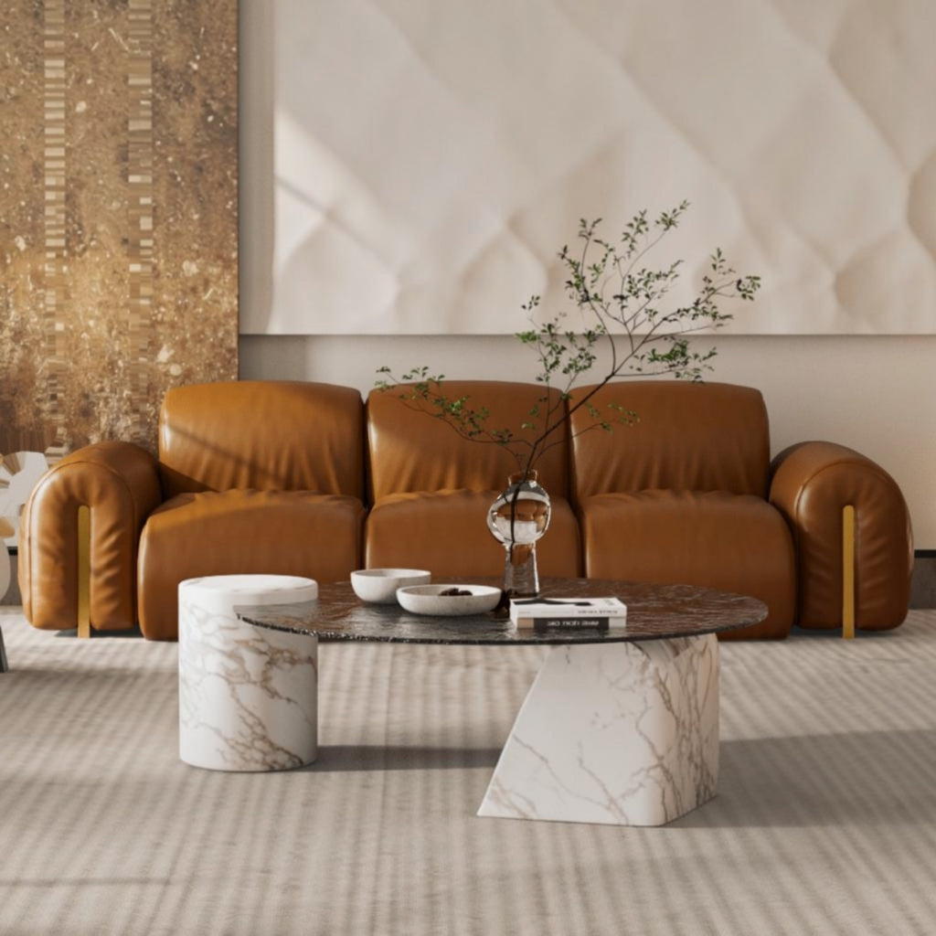A modern living room scene featuring a warm brown leather sofa, a round marble-top coffee table with a white marble pedestal base, two matching marble stools, and a textured beige wall backdrop.