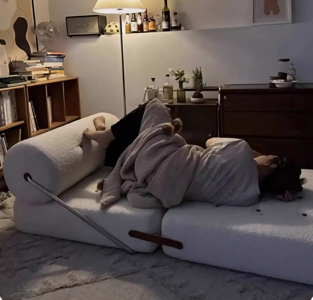 Person lying on a unique sofa with a blanket, surrounded by books and decor in a cozy room.