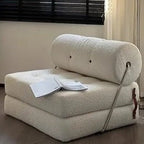 Foldable beige sofa with a pillow and blanket on a wooden floor.
