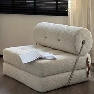 Foldable beige sofa with a pillow and blanket on a wooden floor.